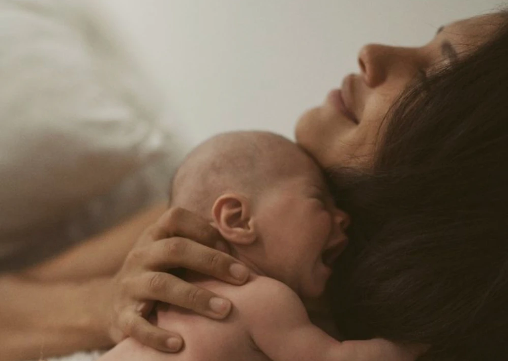 Eine dunkelhaarige Mutter mit ihrem Neugebornen, das sie im Liegen auf sich hält. Die Mutter wirkt freudig, geerdet, glücklich, sicher. Das Baby weint. Sie kann sein Gefühle halten. Das Nervensystem des Babys kann sich über die Mutter regulieren.