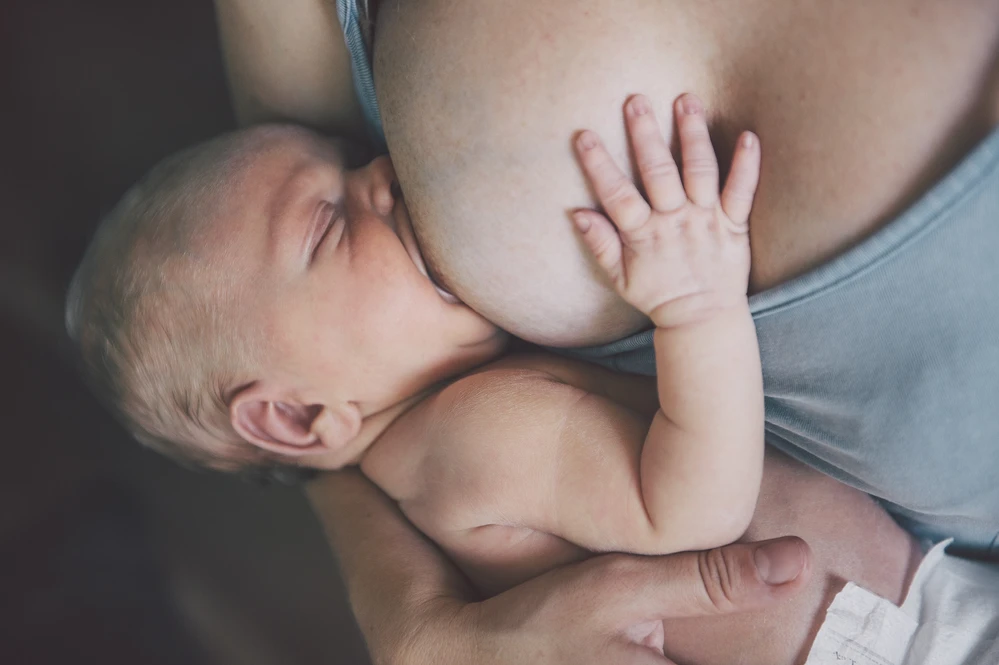 Eine Frau stillt ihr neugeborenes Baby im Wochenbett. Das Baby wirkt müde und entspannt, es hat die Augen zu und sucht Kontakt mit seiner Hand zur Mutter. Die Anlegetechnik ist sehr gut. Gutes Stillmanagement. Viel Brust im Mund.
